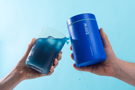 Hands holding a blue supplement container and a glass of blue liquid with a splash between them.